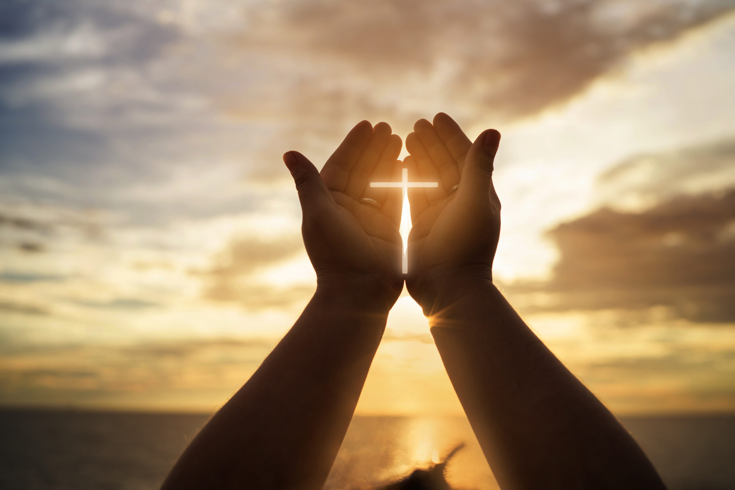 Hands framing a glowing cross against a sunset over the ocean.